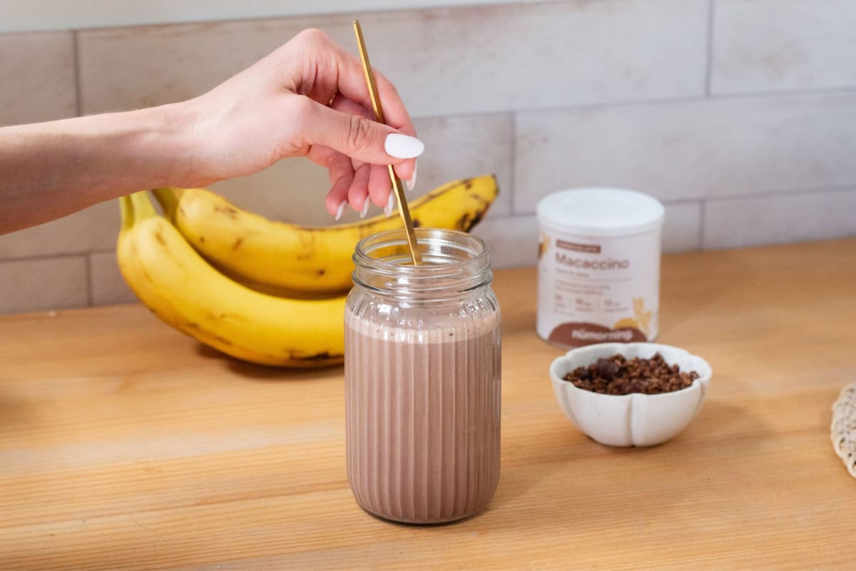 Smoothie végan chocolat dans un verre, avec des bananes et un pot de cacao en poudre en arrière-plan.