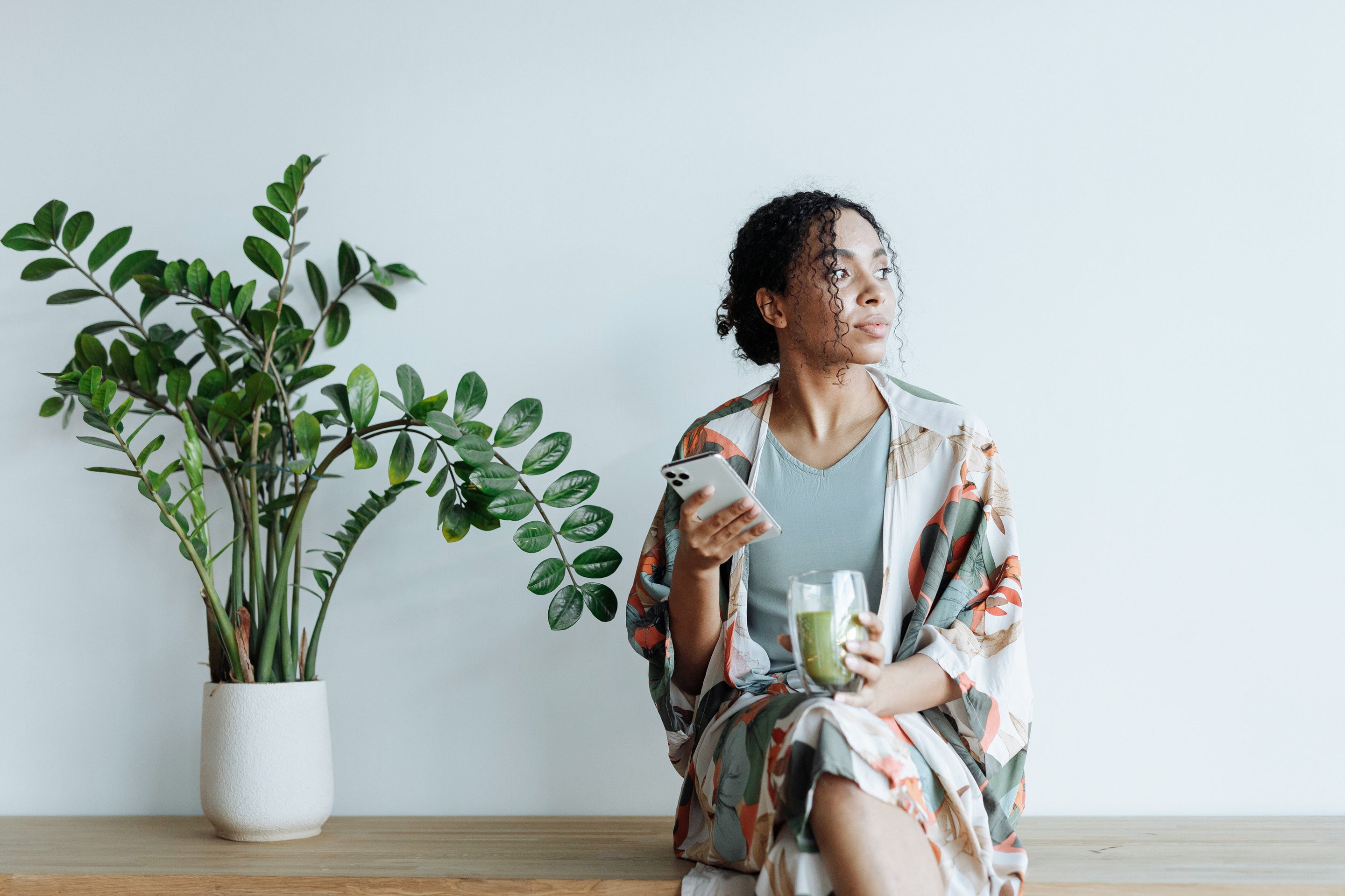 Une femme boit du matcha sur un banc à côté d'une plante en tenant son smartphone